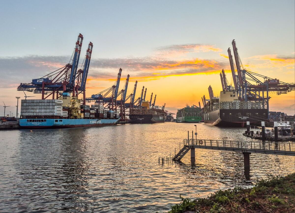 Een brede foto van een haven met meerdere grote containerschepen en imposante havenkranen, tegen een achtergrond van een zonsondergang met oranje en gele tinten aan de horizon. Op de voorgrond is water te zien met een kleine steiger en wat groen aan de oever.