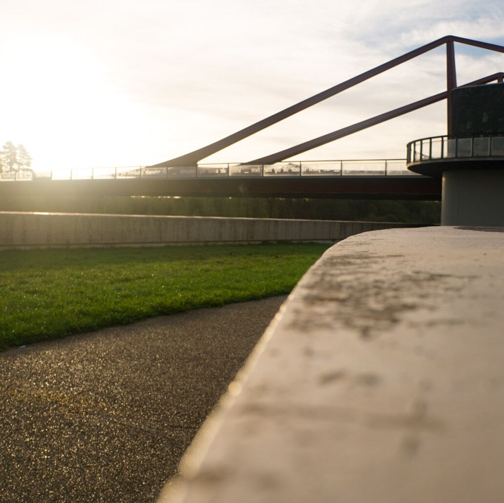 Een moderne brug met rode stalen constructie en een observatieplatform, gezien vanuit een laag perspectief. De zon staat laag aan de horizon en schijnt fel, wat zorgt voor een sterke lichtinval en schaduwen. Op de voorgrond is een betonnen muur, een pad en gras te zien.
