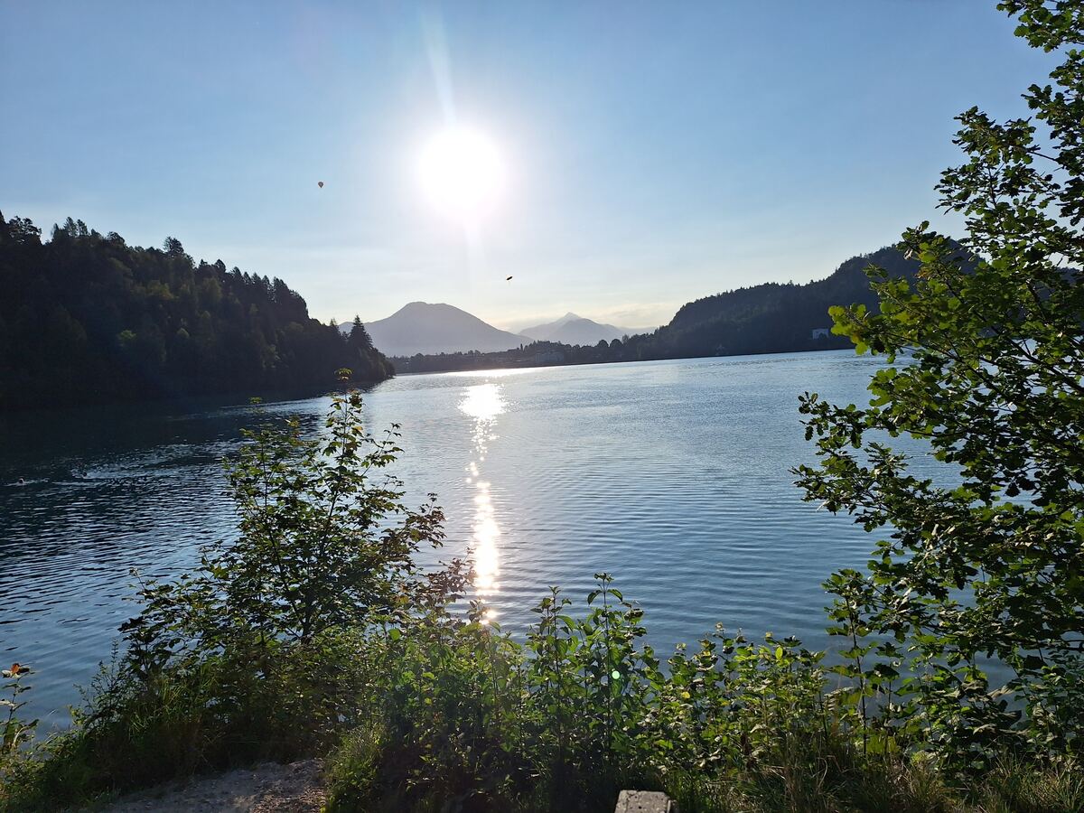 Een breed shot van het meer van Bled met de zon laag aan de horizon, reflecterend op het water. Rondom het meer zijn beboste heuvels en bergen te zien. Op de voorgrond staan enkele groene struiken en bomen. In de verte is een luchtballon zichtbaar.