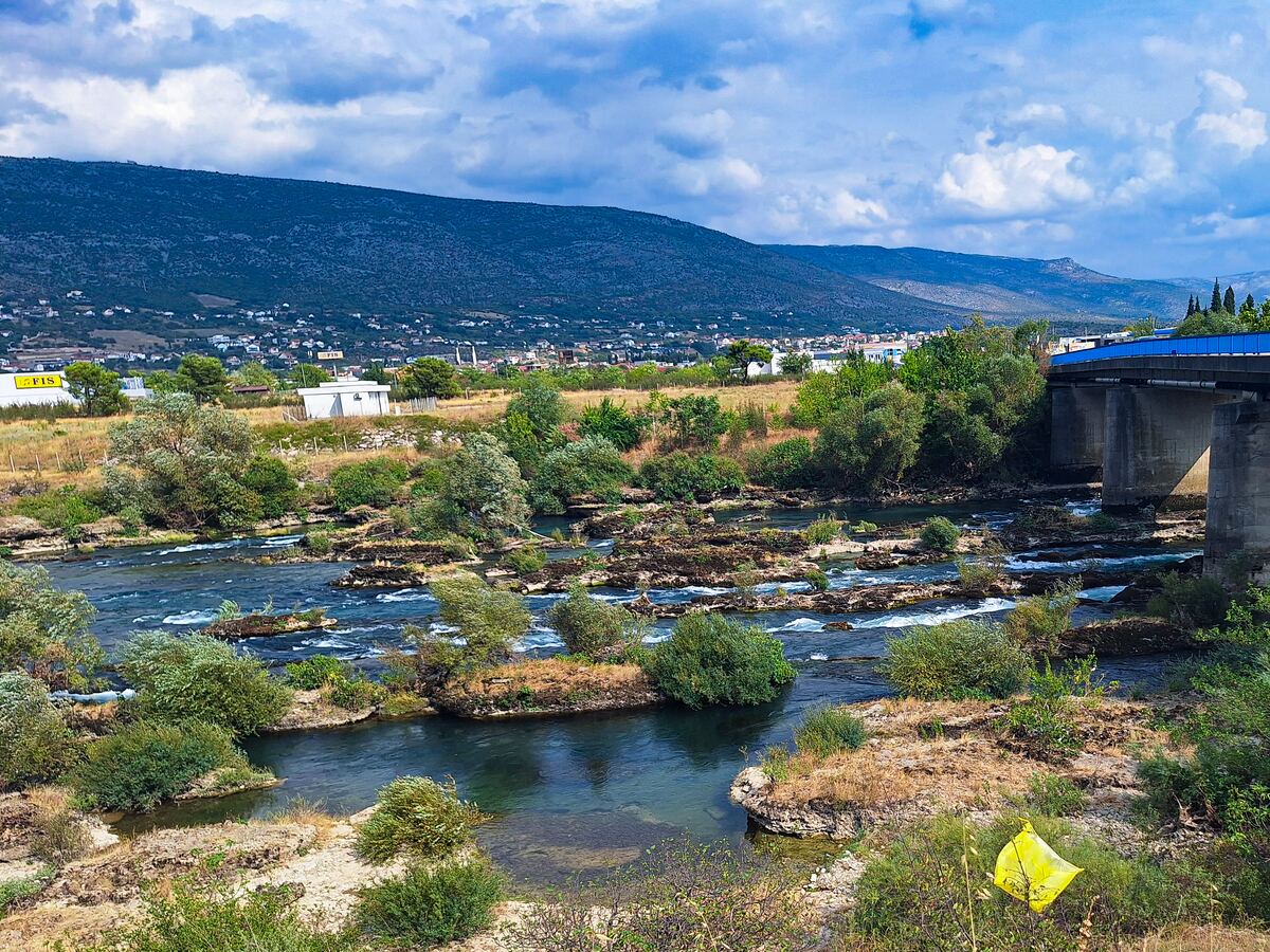 Een brede rivier met veel rotsen en begroeiing in het water stroomt onder een blauwe brug door. Op de achtergrond is een stadje te zien tegen een berghelling onder een bewolkte lucht. Aan de linkerkant staat een geel bord met 'FIS' erop.