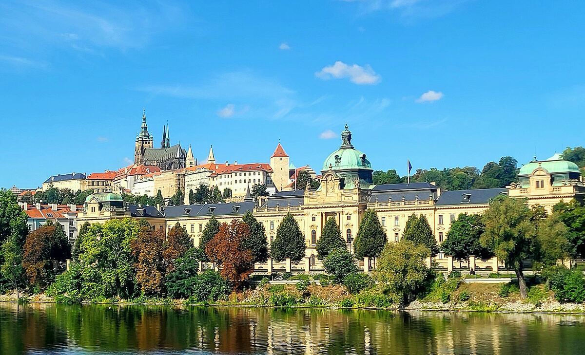 Een breed shot van de Moldau met op de achtergrond de skyline van Praag, inclusief de Sint-Vituskathedraal en andere historische gebouwen. De rivier reflecteert de blauwe lucht en de gebouwen. Aan de oever staan bomen met herfstkleuren.