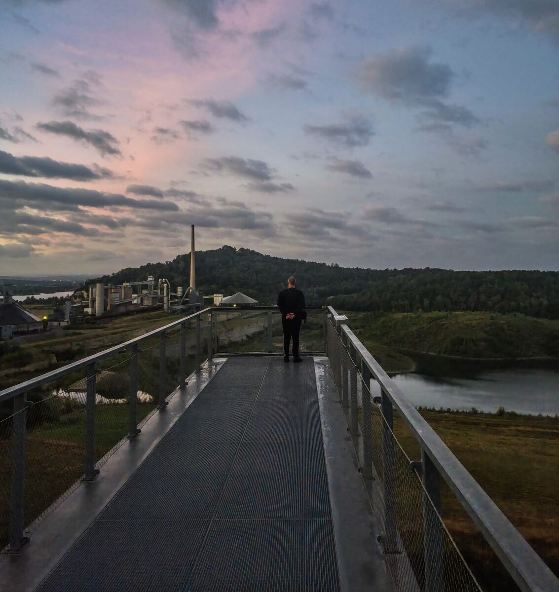 Een persoon staat op een metalen uitkijkplatform en kijkt uit over een industrieel landschap met een fabriek, een schoorsteen, heuvels, water en een deels bewolkte lucht met roze tinten.