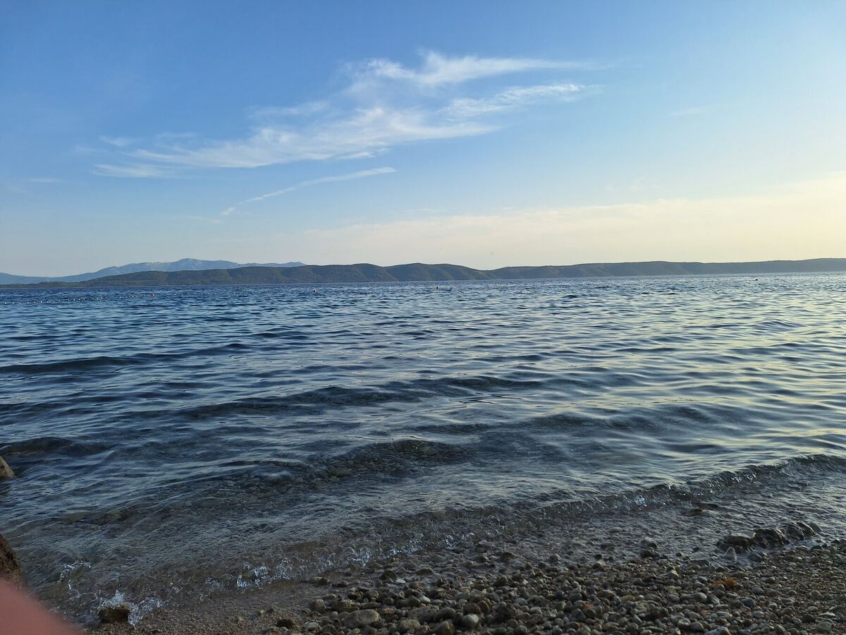 Een foto van een kiezelstrand met kalm water en een heuvelachtig landschap op de achtergrond onder een blauwe lucht met lichte bewolking. De zon staat laag en werpt een zachte gloed over de horizon.