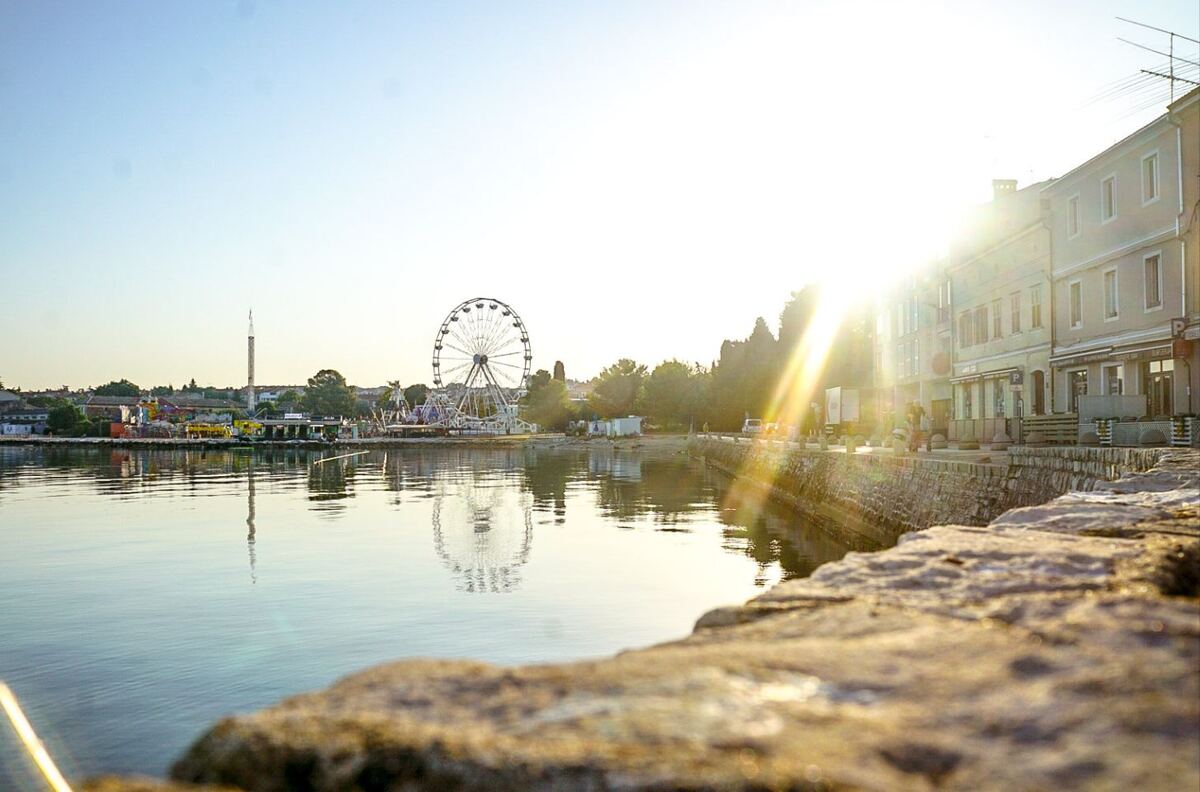 Een brede foto van een waterkant met een kermis aan de overzijde, inclusief een groot reuzenrad. De zon schijnt fel vanaf de rechterbovenhoek en weerkaatst in het water. Aan de rechterkant van de foto zijn gebouwen langs de waterkant te zien, met een stenen muur op de voorgrond.