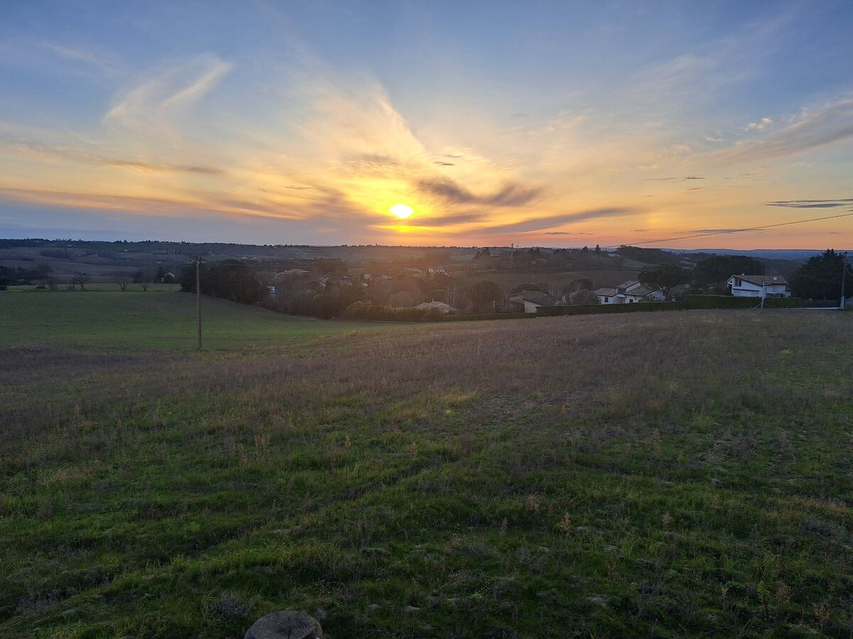 Een weids uitzicht over glooiende velden met op de achtergrond een dorpje en een ondergaande zon die de lucht oranje en geel kleurt. De voorgrond bestaat uit een groen en bruin grasveld.