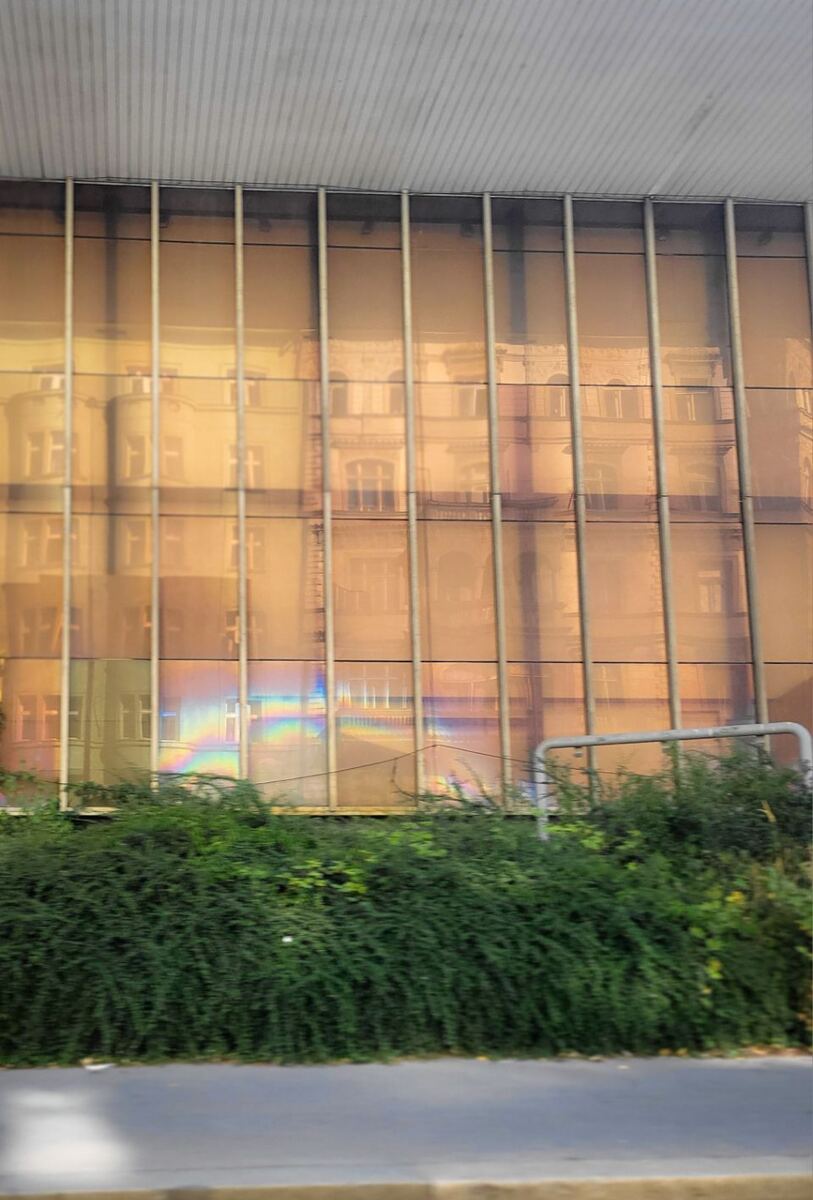 Een verticale foto van een modern gebouw met een goudkleurige, reflecterende gevel. In de gevel is de spiegeling te zien van oudere, klassieke gebouwen met ramen en balkons. Onderaan de gevel is een regenboogeffect zichtbaar. Voor het gebouw staat een rij groene struiken en een deel van een grijs metalen hek. De onderkant van de foto toont een stukje straat en stoep.