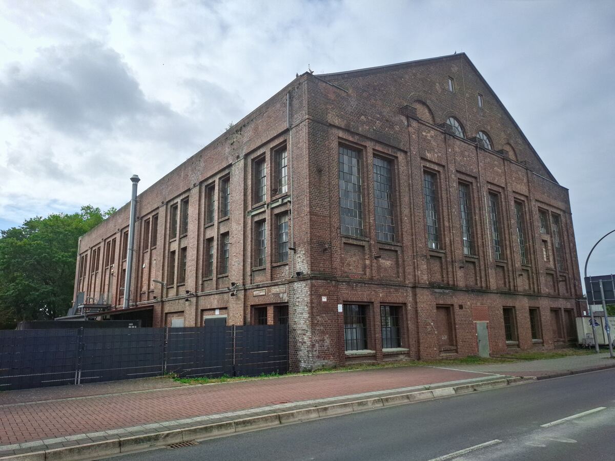 Een groot, oud bakstenen gebouw met veel ramen, gezien vanaf de straat. Het gebouw heeft een industriële uitstraling met een schoorsteen aan de zijkant en een schuin dak. De lucht is bewolkt en er is wat groen van bomen zichtbaar aan de linkerkant.