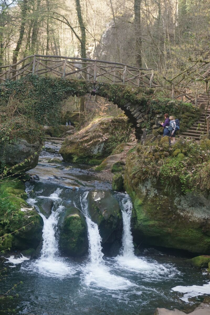 Een stenen boogbrug, deels begroeid met klimop, overspant een beek met kleine watervallen. Twee personen met rugzakken staan rechts van de brug op een rotsachtig pad. De omgeving is bosrijk met veel bomen en rotsen.
