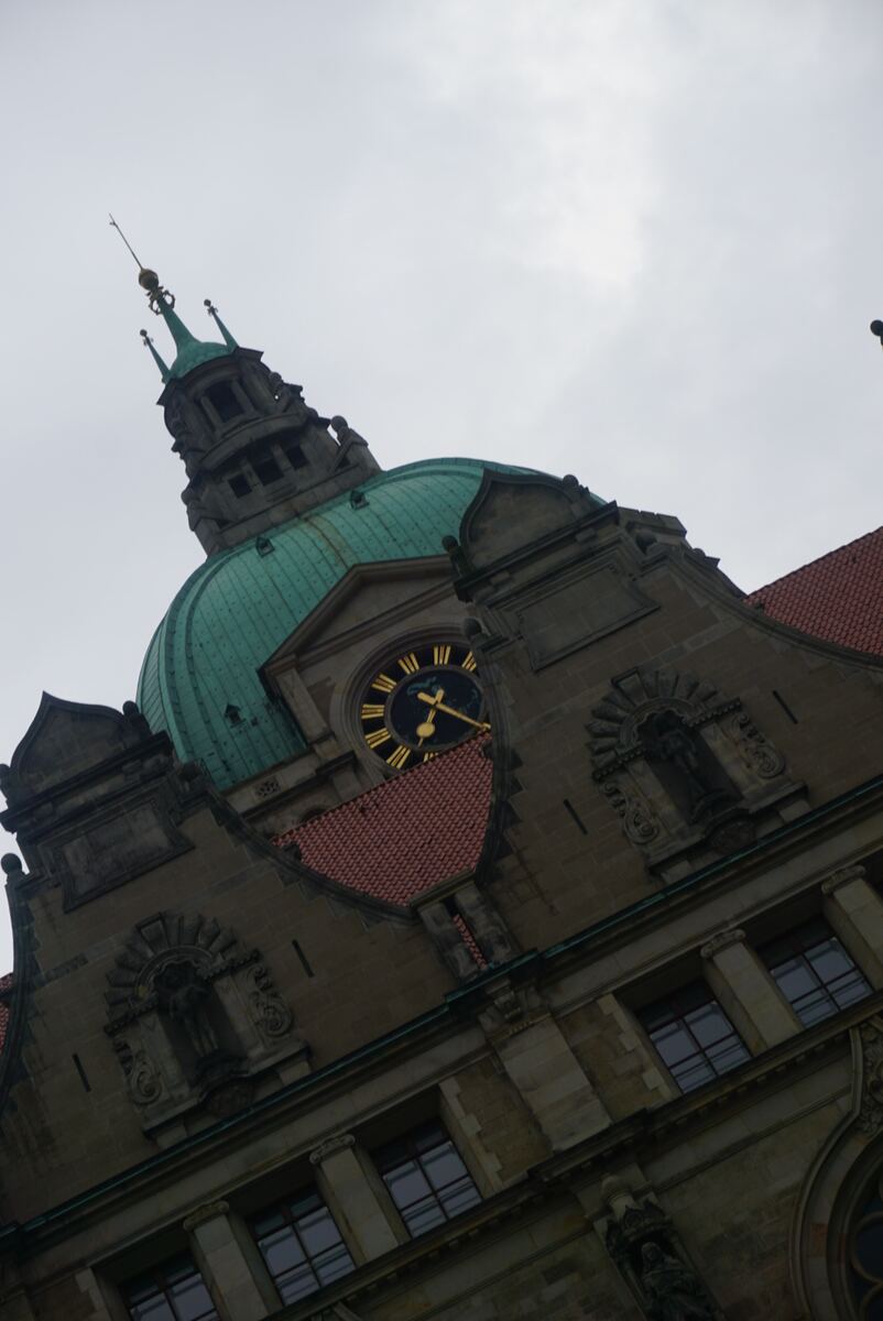Een schuin genomen foto van een klokkentoren met een kopergroene koepel en een wijzerplaat met Romeinse cijfers. De toren is onderdeel van een stenen gebouw met rode dakpannen en gedetailleerde architectuur, waaronder beelden in nissen en versierde raamkozijnen. De lucht is bewolkt en grijs.