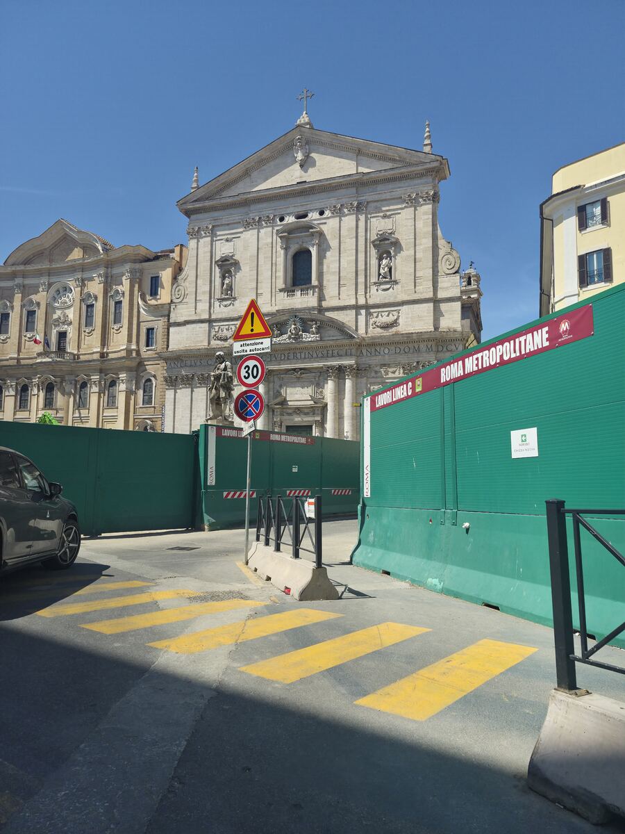 Een barokke kerk met een gedetailleerde gevel staat onder een helderblauwe lucht. Op de voorgrond zijn groene bouwschuttingen geplaatst die de toegang tot de kerk lijken te blokkeren. Op de schuttingen staat 'ROMA METROPOLITANE' en 'LAVORI LINEA C'. Er zijn verkeersborden zichtbaar, waaronder een waarschuwing voor uitrijdende vrachtwagens, een snelheidslimiet van 30 km/u en een parkeerverbod. Een deel van een auto en een zebrapad zijn linksonder in beeld.