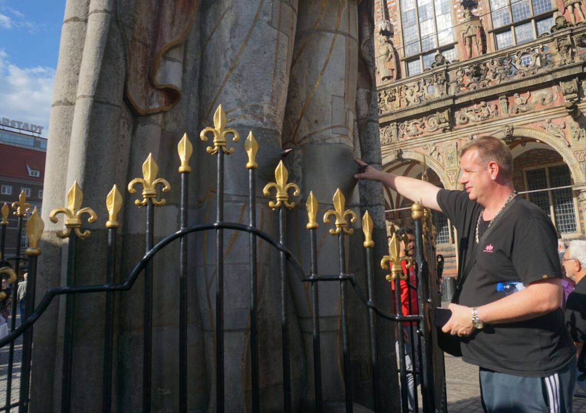 Een man in een zwart T-shirt en een grijze joggingbroek wijst naar een deel van de Roland in Bremen, die deels achter een zwart hek met gouden fleur-de-lis decoraties staat. Op de achtergrond is een deel van het stadhuis van Bremen te zien en een gebouw met de naam 'KARSTADT'.