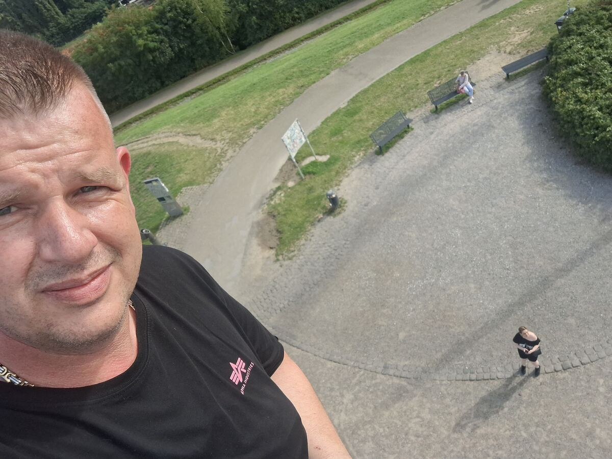 Een man maakt een selfie van bovenaf, met op de achtergrond een parklandschap met paden, bankjes en groen. Een vrouw staat beneden op het pad en kijkt omhoog. Verderop zitten twee mensen op een bankje.