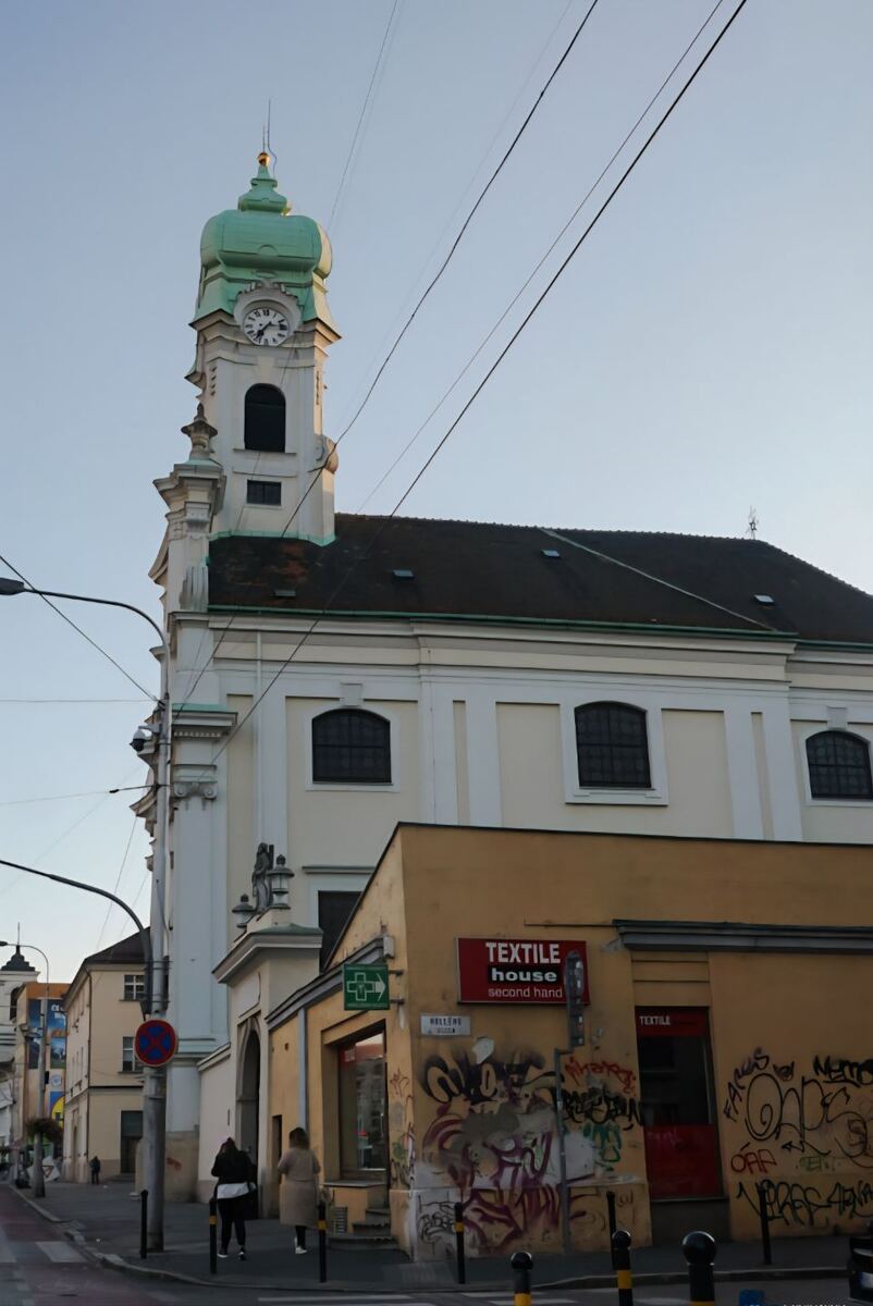 Een foto van een barokke kerk met een groene koepel en klokkentoren, gefotografeerd vanuit een laag perspectief. Op de voorgrond is een geel gebouw met graffiti te zien, met een uithangbord 'TEXTILE house second hand'. Er lopen twee personen op de stoep en er zijn bovengrondse elektriciteitskabels zichtbaar tegen een helderblauwe lucht.