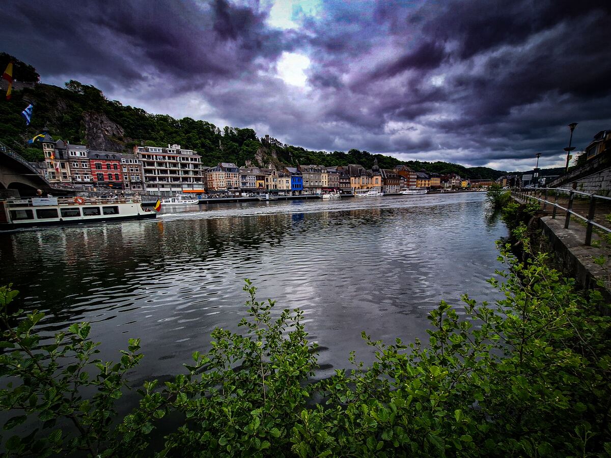 Een breed overzicht van de Maas in Dinant, België, met een donkere, bewolkte lucht erboven. Aan de linkerkant van de rivier liggen gebouwen langs de oever en op de achtergrond is een beboste heuvel te zien. Op de rivier varen enkele boten, waaronder een rondvaartboot. Op de voorgrond zijn groene struiken zichtbaar en rechts loopt een kade met een reling.