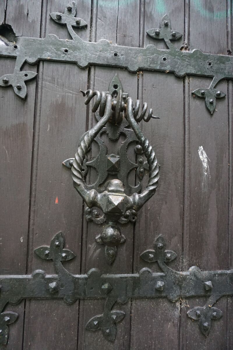 Een close-up van een donkerbruine houten deur met een grote, sierlijke metalen deurklopper en bijpassend beslag. Het beslag bestaat uit horizontale banden met decoratieve, bladvormige elementen.