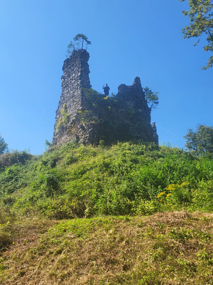 Een lage camerahoek toont de ruïne van een stenen kasteel op een met groen begroeide heuvel onder een helderblauwe lucht. Een persoon staat op de ruïne en zwaait naar de camera. De ruïne is deels overwoekerd met klimop en er groeien kleine bomen op de top.