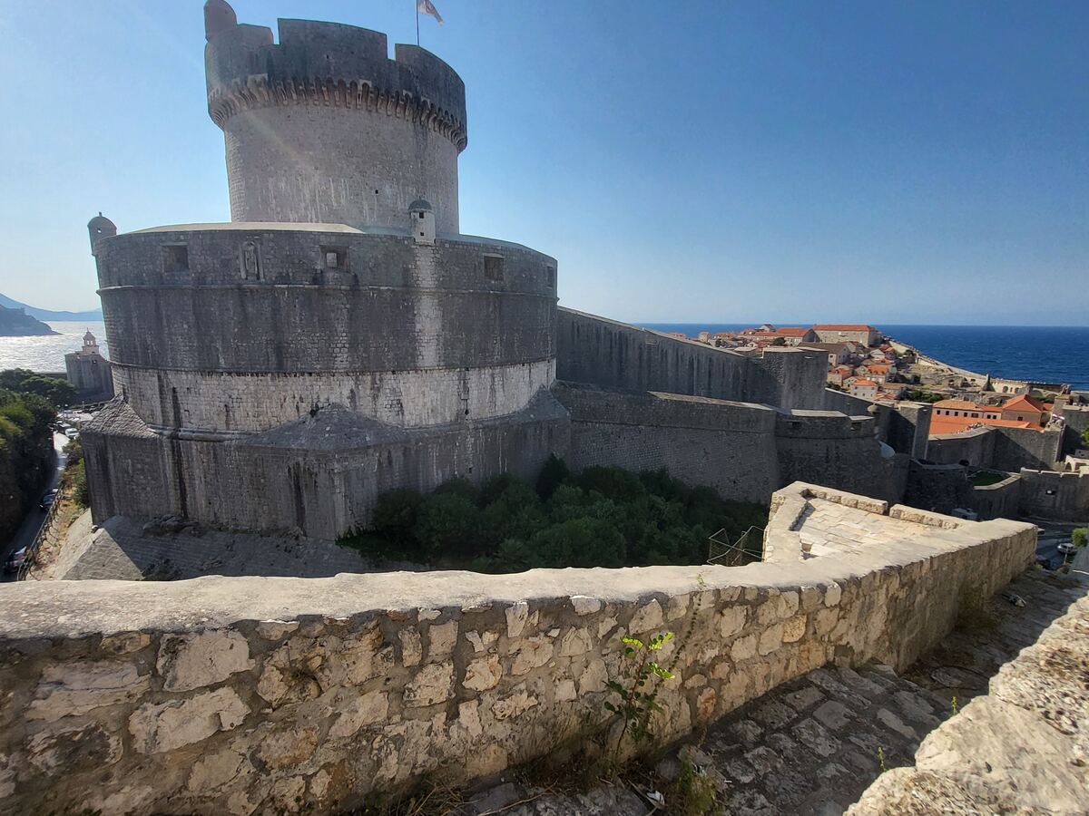 Een foto van de imposante stadsmuren van Dubrovnik, met een grote ronde toren aan de linkerkant. Rechts daarvan strekken de muren zich uit langs de kustlijn, met daarboven de rode daken van de oude stad en de helderblauwe Adriatische Zee. Op de toren wappert een vlag en op de voorgrond is een stenen muur te zien.