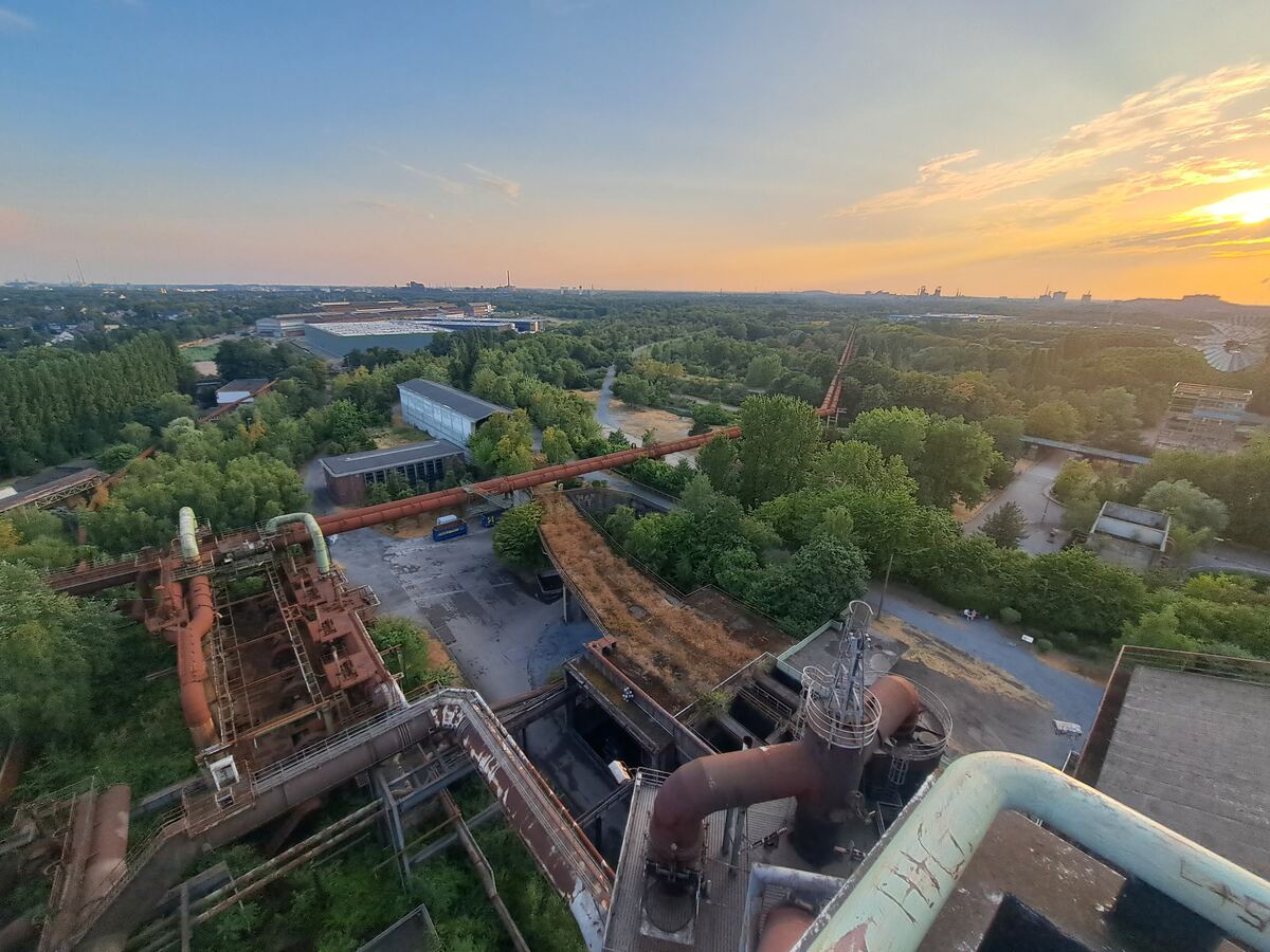Een weids uitzicht over een voormalig industrieel complex, gezien vanaf een hoog punt. Grote roestige pijpleidingen en metalen structuren domineren de voorgrond, omgeven door weelderig groen en bomen. In de verte zijn meer industriële gebouwen en structuren zichtbaar. De lucht toont de kleuren van de zonsondergang, variërend van blauw naar oranje en geel, met enkele wolken. Er zijn paden te zien die door de begroeiing slingeren.