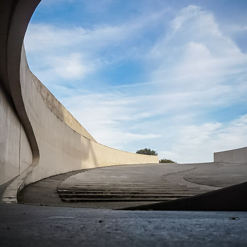 Een foto van een modern architectonisch bouwwerk met een lange, gebogen betonnen muur en een brede trap die omhoog loopt. De muur heeft een golvende vorm aan de linkerkant. Boven de constructie is een helderblauwe lucht met enkele witte wolken zichtbaar. Een paar bomen zijn te zien aan de horizon.