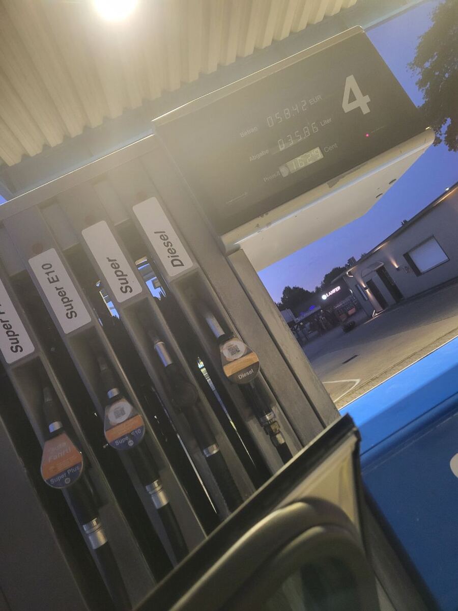 Een close-up van een brandstofpomp bij schemering, met de display die het bedrag van 58,42 EUR en 35,86 liter toont, en een prijs van 1,629 cent per liter. De pomp heeft verschillende brandstofsoorten zoals Super E10, Super en Diesel. Op de achtergrond is een gebouw van een tankstation te zien met een verlicht bord 'LANFER'.