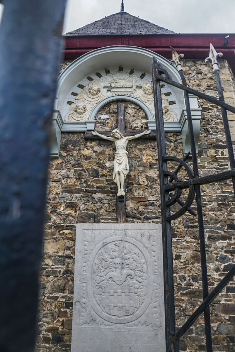 Een close-up van een stenen muur met een kruisbeeld van Jezus Christus. Boven het kruisbeeld is een boogvormige nis met engelenfiguren en een lam. Onder het kruisbeeld staat een grote, rechthoekige steen met een wapenschild. Rechts is een deel van een zwart metalen hek zichtbaar.