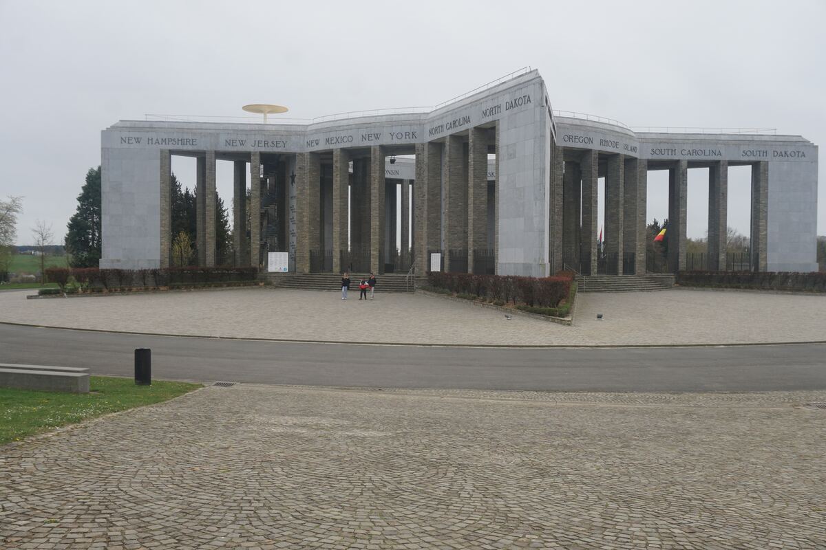 Een groothoekopname van het Mardasson Memorial, een stervormig monument met zuilen en de namen van Amerikaanse staten erop. Er staan enkele mensen bij de ingang van het monument. De lucht is bewolkt en de omgeving bestaat uit gras en een geplaveid plein.