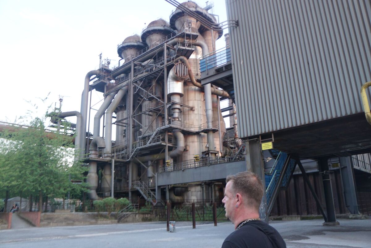 Een groothoekopname van een groot industrieel complex met meerdere hoge, ronde constructies, pijpen en stalen platforms. Aan de rechterkant is een deel van een gebouw met verticale lamellen zichtbaar. Links staan enkele bomen. Op de voorgrond is de achterkant van een persoon te zien, kijkend naar de constructie. Verder naar links, in de verte, zit nog een persoon.