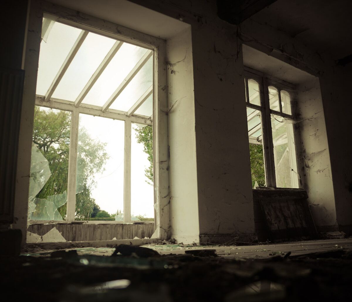 Een interieur van een verlaten gebouw met twee grote ramen. Het linker raam is grotendeels gebroken, met scherven op de vensterbank en de vloer. Door de ramen is groen gebladerte van bomen zichtbaar. De muren zijn verweerd en de vloer is bedekt met puin en stof.