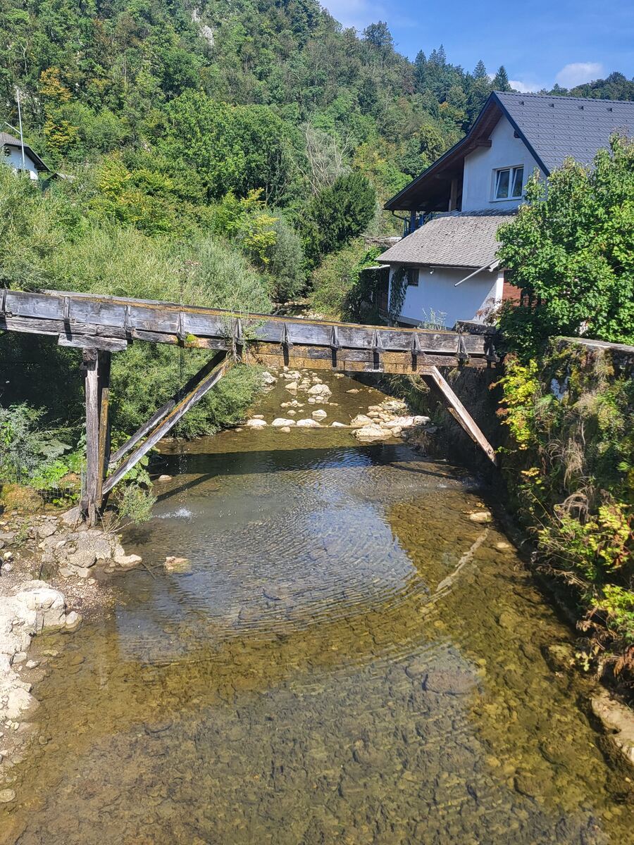 Een houten constructie, mogelijk een oude waterloop of aquaduct, overspant een ondiepe rivier met een rotsachtige bedding. Aan de rechteroever staat een wit huis met een donker dak, omgeven door groene begroeiing. Op de achtergrond is een beboste heuvel te zien onder een blauwe lucht met enkele wolken.