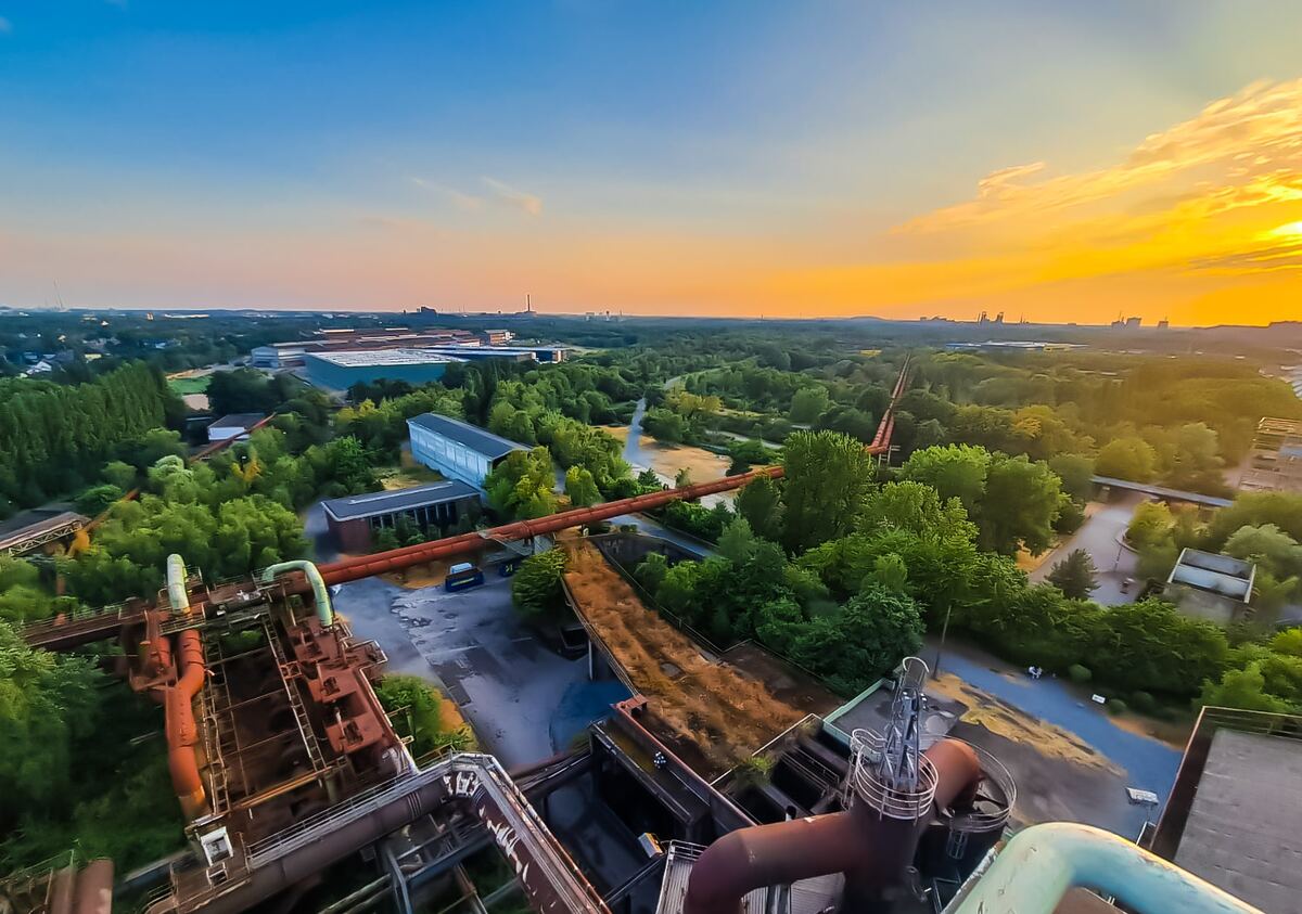Een weids uitzicht over het Landschaftspark Duisburg-Nord, genomen vanaf een hoger punt. Op de voorgrond zijn roestbruine industriële constructies en pijpleidingen te zien. Het midden- en achterplan wordt gedomineerd door uitgestrekte groene bomen en struiken, afgewisseld met enkele gebouwen en paden. Aan de horizon strekt zich een oranje-gele lucht uit, overgaand in blauw bovenaan, wat duidt op de schemering.