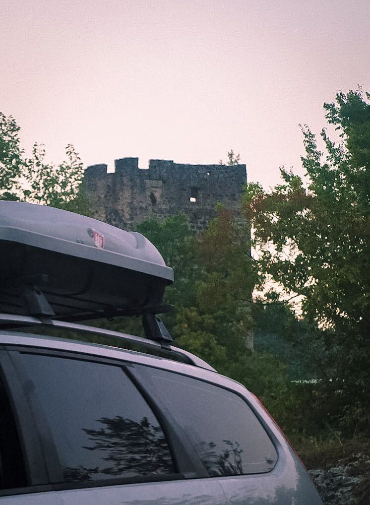Een foto van de achterkant van een zilverkleurige auto met een dakkoffer, met op de achtergrond een oude stenen ruïne die deels verscholen is achter bomen. De lucht is lichtroze getint, wat duidt op zonsopgang of zonsondergang.
