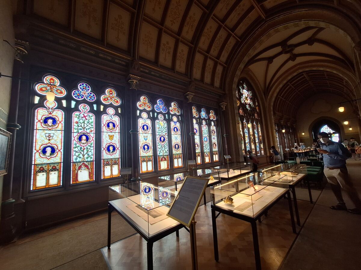 Een interieur van een gebouw met hoge, gedetailleerde glas-in-loodramen die veel licht doorlaten. Er staan vitrines met tentoonstellingsstukken in de ruimte en enkele bezoekers zijn zichtbaar. Het plafond is gewelfd en versierd.