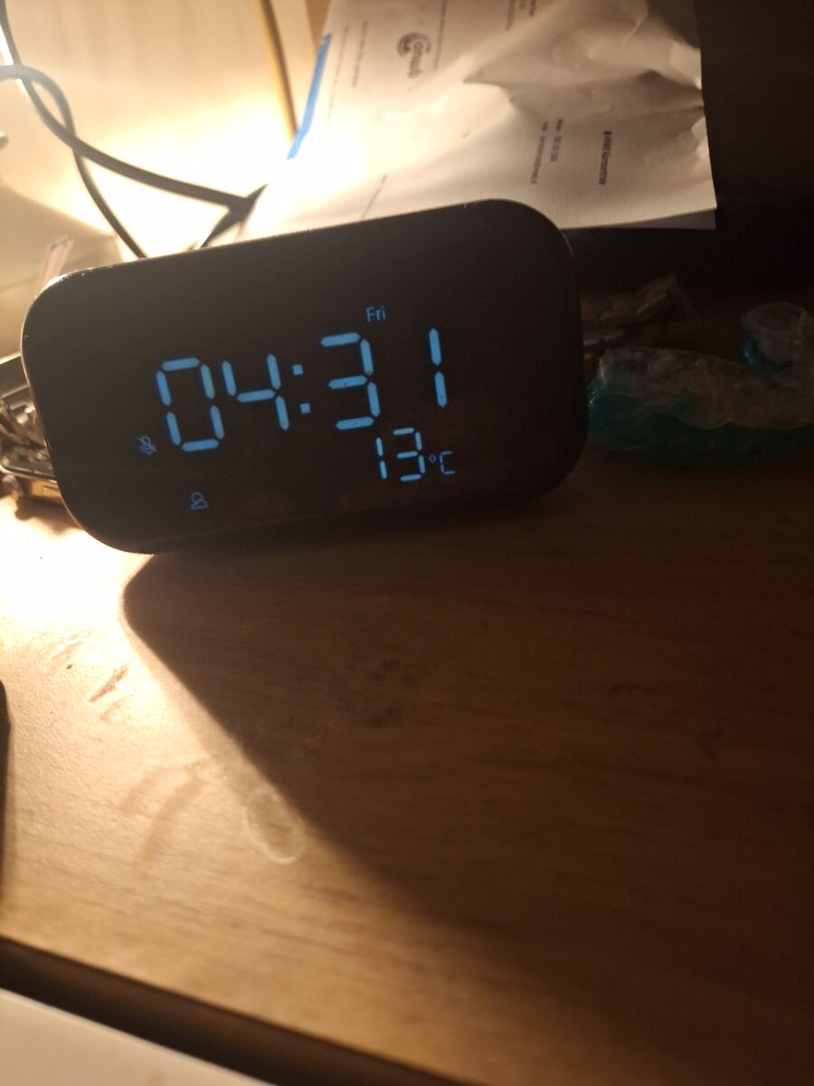 Een digitale wekker op een houten nachtkastje toont 04:31 uur en 13°C. Er is een zwakke lichtbron zichtbaar en wat papieren op de achtergrond.