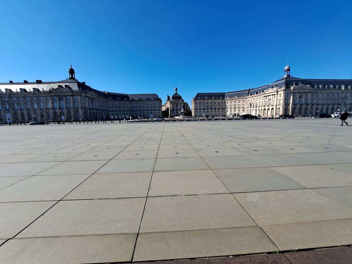 Een weids, open plein met grote grijze tegels onder een strakblauwe lucht. Aan weerszijden van het plein staan imposante klassieke gebouwen met donkere daken en veel ramen. In het midden van het plein, verder weg, is een fontein met een gebouw erachter zichtbaar. Enkele mensen lopen verspreid over het plein en langs de gebouwen.