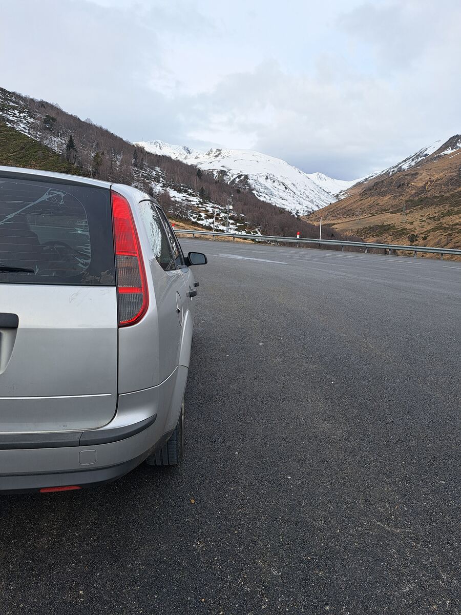 De achterkant van een zilverkleurige Ford Focus stationwagon staat geparkeerd langs een geasfalteerde weg in een berglandschap. De weg loopt een vallei in. Links zijn besneeuwde hellingen te zien met kale bomen, terwijl rechts de hellingen bruiner zijn met minder sneeuw. Op de achtergrond verheffen zich besneeuwde bergtoppen onder een bewolkte lucht. Een vangrail loopt langs de weg.
