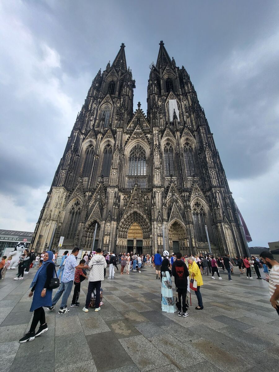 Een groothoekopname van de Dom van Keulen, gezien vanaf de voorzijde, met de twee hoge torens die de bewolkte lucht in steken. Op het plein voor de kathedraal zijn veel mensen te zien die rondlopen en de Dom bekijken.