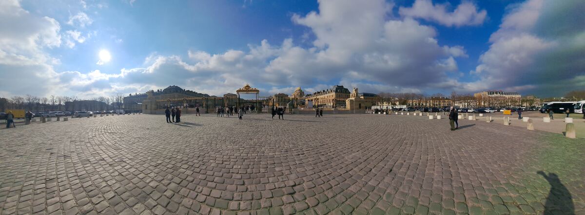 Een panoramische foto van het Place d'Armes voor het Paleis van Versailles, met de gouden poorten en gebouwen van het paleis onder een deels bewolkte lucht. Er zijn mensen en geparkeerde auto's zichtbaar op het plein.
