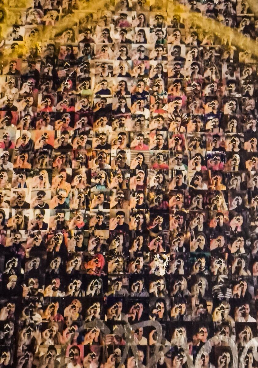 Een close-up van een mozaïekkunstwerk dat bestaat uit honderden kleine foto's van mensen die met hun handen een hartje vormen voor hun gezicht. De foto's zijn in een raster geplaatst en vormen samen een groter, abstract patroon.