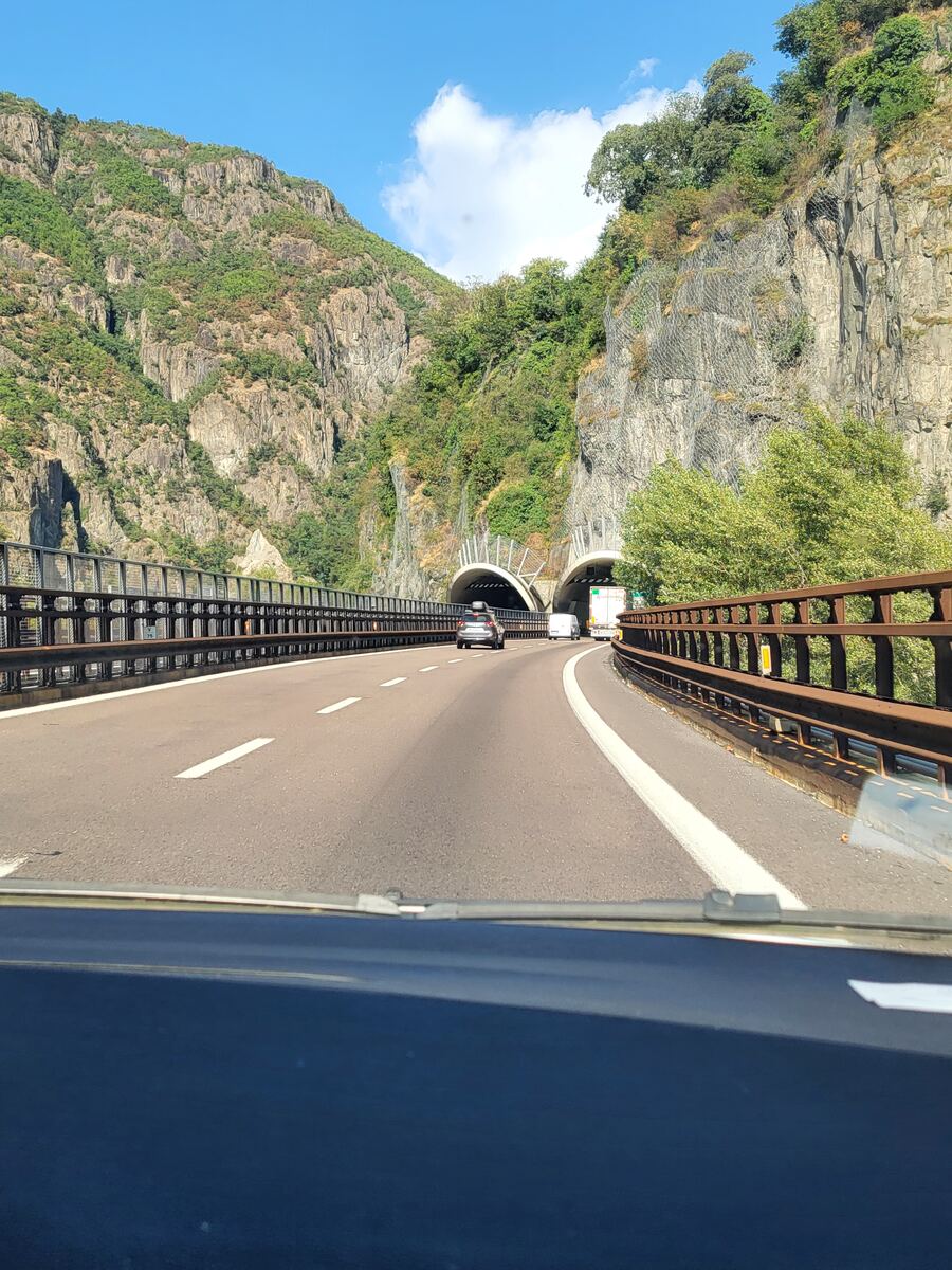 We rijden op een snelweg met twee rijstroken in elke richting, omringd door hoge, rotsachtige bergen met groene begroeiing. Voor ons zijn twee tunnels zichtbaar. De weg buigt licht naar rechts en er rijden enkele auto's en een vrachtwagen voor ons. De zon schijnt en de lucht is blauw met een paar witte wolken.