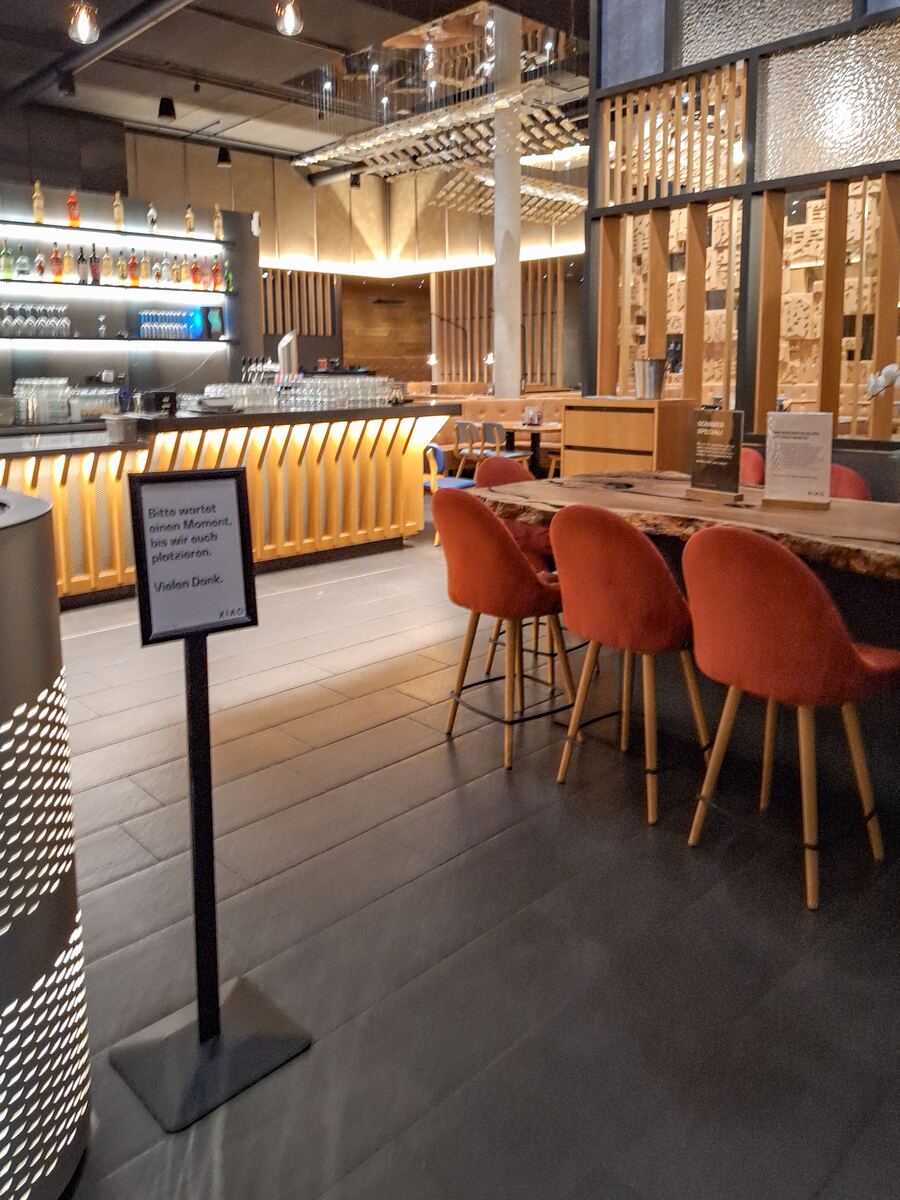 Een modern restaurantinterieur met een lange bar aan de linkerkant, verlicht met warm geel licht. Er staan flessen drank op planken achter de bar. Op de voorgrond staan vier oranje stoelen aan een houten tafel. Een bordje met Duitse tekst vraagt bezoekers te wachten tot ze geplaatst worden.