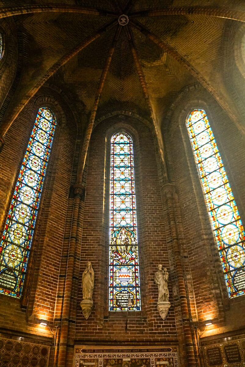 Een interieurfoto van een kerk of kathedraal met hoge bakstenen muren en gewelfde plafonds. Drie grote, verticale glas-in-loodramen met gedetailleerde, kleurrijke patronen zijn zichtbaar, die licht doorlaten. Tussen de ramen hangen twee beelden van heiligen aan de muur. De architectuur is gotisch of neogotisch van stijl.