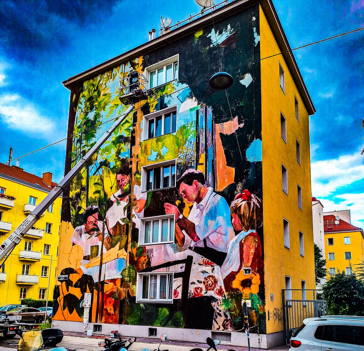 Een gevel van een gebouw is beschilderd met een groot, kleurrijk kunstwerk dat mensen in een bosrijke omgeving uitbeeldt. Een hoogwerker met een persoon erin is zichtbaar aan de linkerkant van het gebouw, bezig met werkzaamheden aan de muurschildering. De lucht is helderblauw met enkele wolken. Rechts van het gebouw staat een witte auto geparkeerd en op de voorgrond zijn scooters en fietsen te zien.