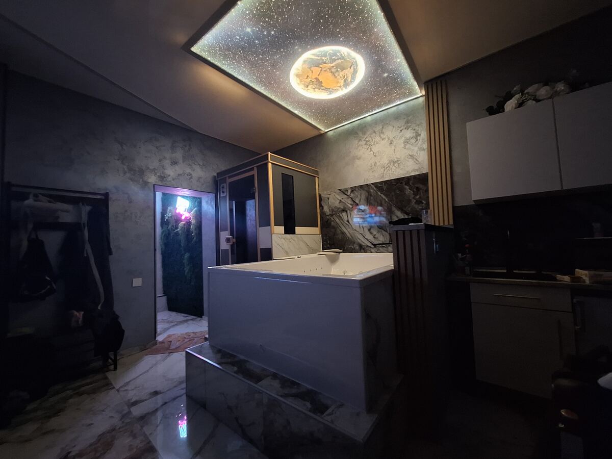 Een sfeervolle badkamer met een groot wit bubbelbad in het midden. Het plafond heeft een verlichte afbeelding van een sterrenhemel met een planeet. De muren zijn deels afgewerkt met donker marmer en deels met een grijze, gestructureerde verf. Rechts is een deel van een keukenblok zichtbaar. Links is een open deur met daarachter een wand bedekt met groen, verlicht door roze neonlicht.