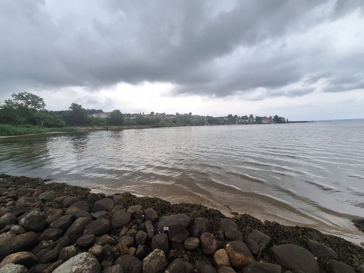 Een bewolkte dag aan het water, met op de voorgrond een rotsachtige oever en op de achtergrond een rij huizen langs de kustlijn. Het water is licht golvend en de lucht is grijs.