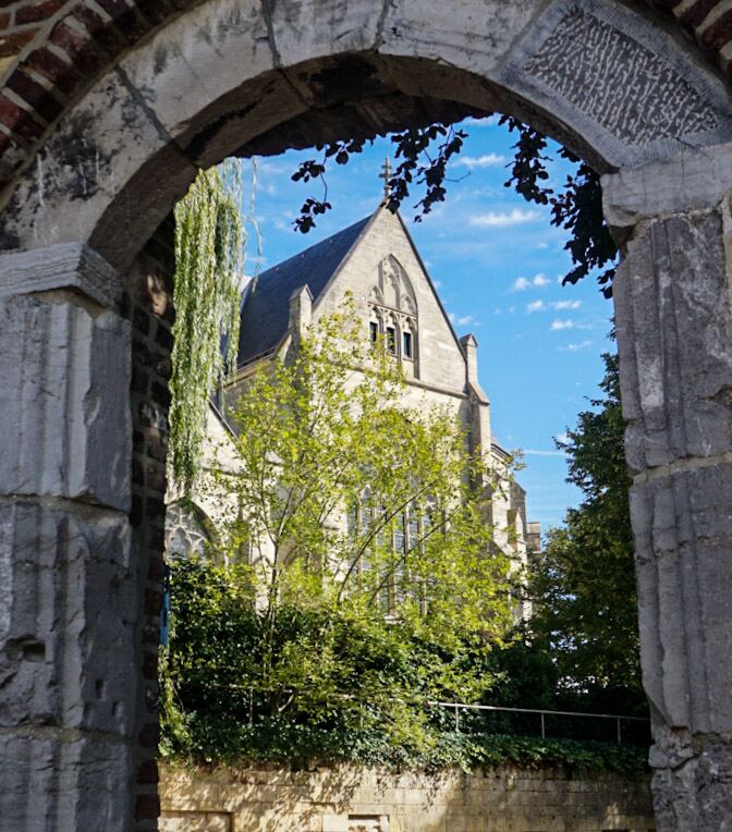Een foto genomen door een oude stenen boog, met op de achtergrond een lichtgekleurd kerkgebouw met een puntig dak en een kruis op de top. Rondom het gebouw is veel groen te zien, waaronder bomen en struiken, en de lucht is helderblauw met enkele witte wolken.