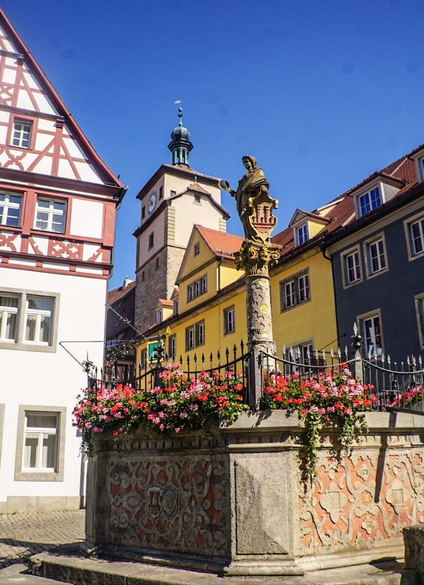 Een stadsgezicht met een stenen fontein versierd met rode en roze bloemen op de voorgrond. Achter de fontein staan gebouwen met traditionele Duitse architectuur, waaronder een vakwerkhuis en een gele gevel. Op de achtergrond is een klokkentoren met een groene koepel te zien onder een helderblauwe lucht.