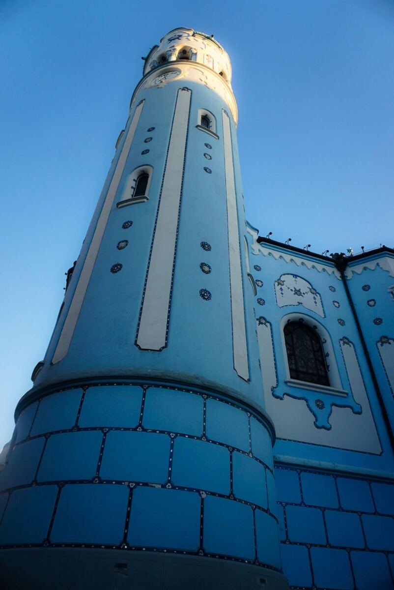 Een laag camerastandpunt van de blauwe Sint-Elisabethkerk in Bratislava, met de toren en een deel van de gevel zichtbaar tegen een helderblauwe lucht. De kerk is versierd met blauwe en witte tegels en details.