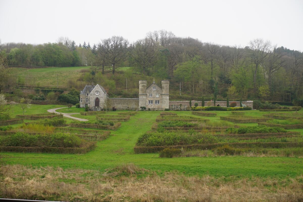 Een stenen gebouw met twee torens en een kleiner bijgebouw, omgeven door een muur en een tuin met lage heggen, gelegen in een heuvelachtig, groen landschap met bomen onder een grijze lucht.
