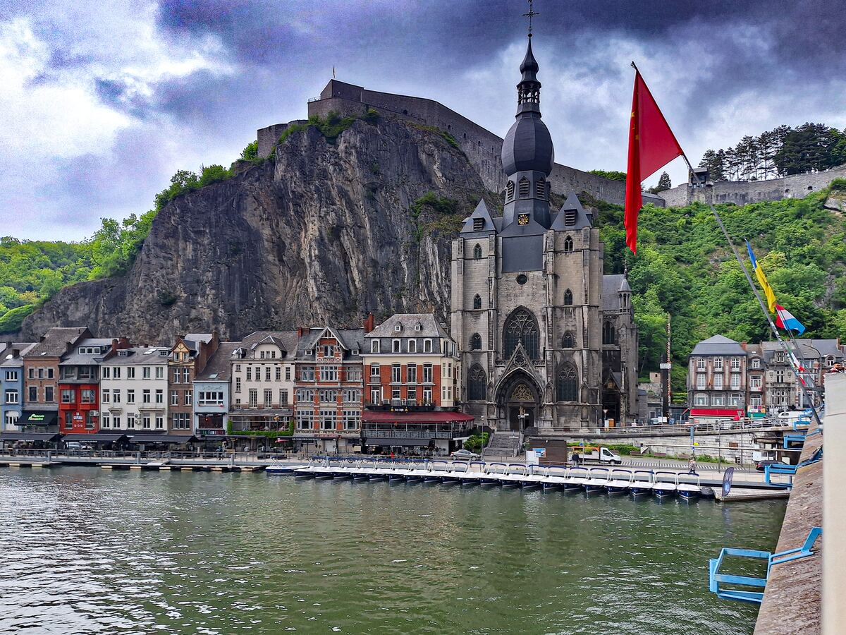 Een brede rivier met aan de overkant een rij gebouwen, een grote kerk en een steile rotswand met daarop een fort. Er hangt een grote rode vlag met gele sterren en een kleinere vlag met blauw, geel en rood.