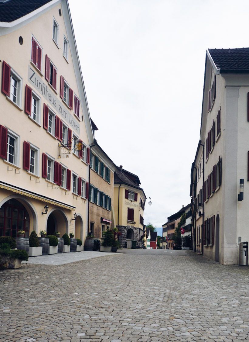 Een geplaveide straat in een Europees stadje met aan weerszijden gebouwen. Links staat een lichtgeel gebouw met rode luiken en de tekst 'Zunfthaus zum Lowen'. Rechts staan ook gebouwen met luiken. De straat loopt licht omhoog en er is een bewolkte lucht zichtbaar.